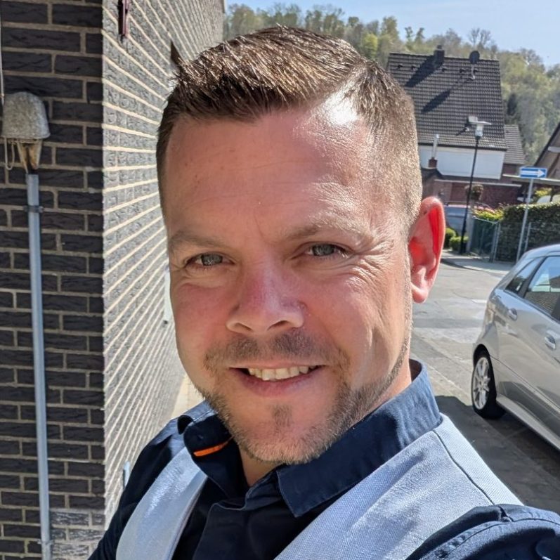 Mann mit kurzem Haar und lächelndem Gesicht in hellem Oberteil vor Häusern.