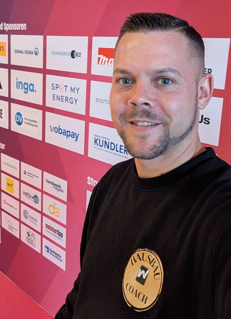 Ein Mann mit kurzem Haar lächelt vor einer Wand mit Sponsorenlogos.