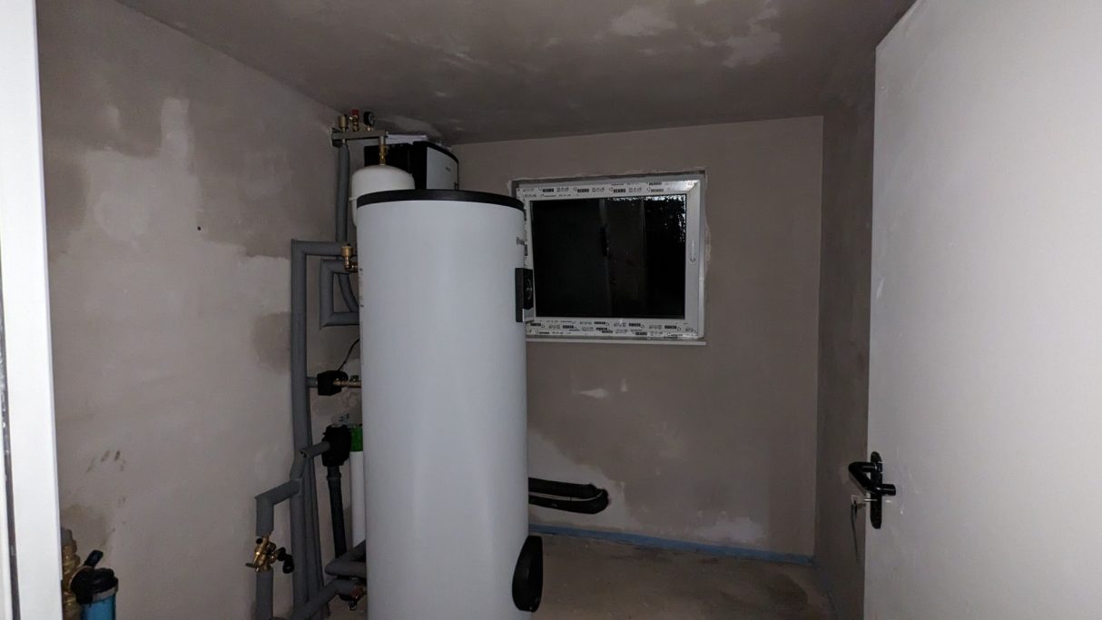 Ein Kellerraum mit einem großen weißen Boiler in der Ecke. An der Wand sind diverse Rohrleitungen zu sehen. Das Fenster ist klein.